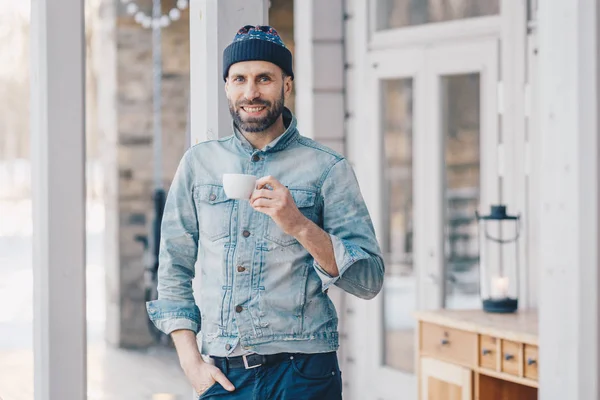 Portrait of attractive stylish bearded male with happy look, wears fashionable denim jacket, hat and trousers, holds mug of hot beverage, recreats at home after hard working day, smiles pleasantly — Stock Photo, Image