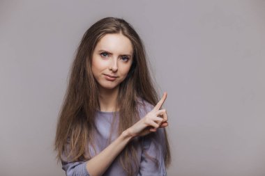 Boş kopya alanı gri duvar, noktalarda esmer uzun saçlı sevimli genç kadın modeli ciddi ifade ile bir şey advertizes, dikkatinizi çekiyor. Reklam ve insanlar kavramı