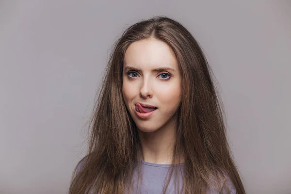 Shot of beautiful brunette female licks lips with tongue, has pure healthy skin, dressed casually, isolated over grey background. Lovely woman shows tongue, poses indoor. Facial expressions. — Stock Photo, Image