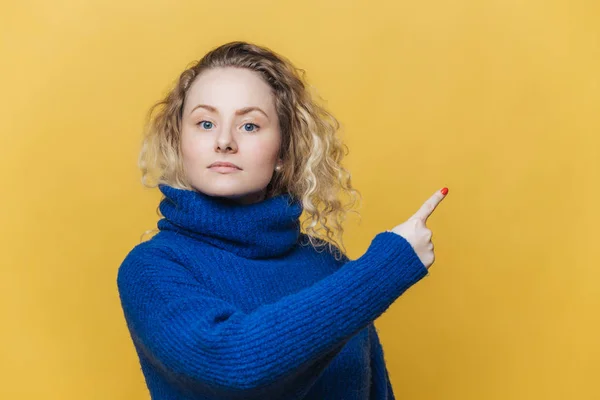 To sprawdzić. Poważne blondynka młoda kobieta z Kręcone blond włosy, ubrany w jasny niebieski sweter, wskazuje na puste miejsce na tekst promocyjny lub reklamowym treści. Pusta ściana żółty — Zdjęcie stockowe