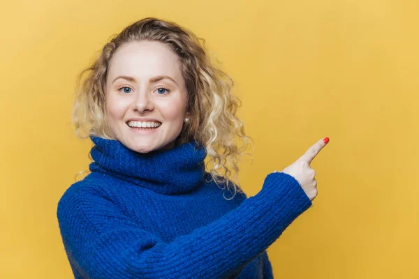 Szczęśliwy, Piękna blondynka młoda kobieta ma przyjemność wyrażenie, nosi na co dzień niebieski sweter, wskazuje na puste miejsce na żółty studio puste ściany, advertizes coś z wielką przyjemnością. Reklama — Zdjęcie stockowe