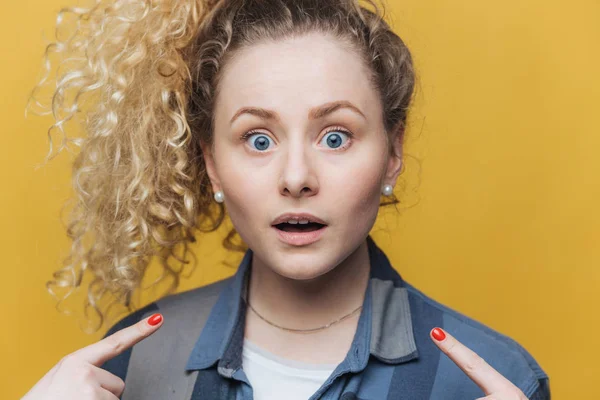 Horizontal shot of pleasant looking young curly woman indicates with fore fingers at herself, being shocked to recieve failure, dressed casually, has amazed expression, isolated over yellow wall