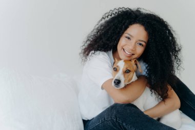 Afro-Kircut 'lu çekici genç bir kadının fotoğrafı, aşk köpeğiyle kucaklaşması, evcil hayvanla ilgilenmesi, nazikçe gülümsemesi, günlük kıyafetler giymesi, beyaz arka planda izole edilmesi, yatağa oturması, tanıtımın için yer kopyalaması.