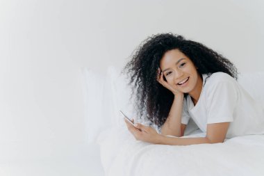 Neşeli, rahat, kıvırcık kıvırcık, genç Afrikalı Amerikalı kadın evde boş vakitlerini geçiriyor, modern cep telefonuyla yatakta uzanıyor, yatak odasında telefon kullanıyor, sevgilisine mesaj atıyor, tembel bir sabah geçiriyor, iyi vakit geçiriyor.