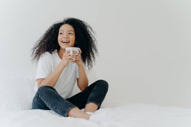 Lazy morning and bedtime concept. Positive Afro woman holds mug of hot tea or coffee, sits in bed, looks gladfully aside, enjoys good weather, wears comfortable clothes, white wall in background