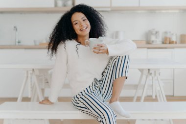 Beautiful young female with Afro haircut, poses on white bench indoor, dressed in stylish clothing, enjoys aromatic beverage, enjoys breakfast at home, poses against blurred kitchen interior