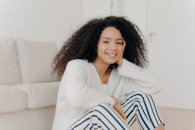 Rahatlamış, kıvırcık saçlı, kameraya nazikçe gülümseyen beyaz kazak ve çizgili pantolon giyen, modern dairenin oturma odasındaki kanepenin yanında oturan Afro-Amerikan kadının fotoğrafı.