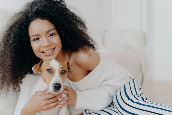 Image de belle femme à la peau foncée porte du maquillage, a les cheveux bouclés touffus, sourit agréablement, câlins chien, habillé dans des vêtements à la mode, bénéficie moment doux avec chiot, être à la maison ensemble — Photo