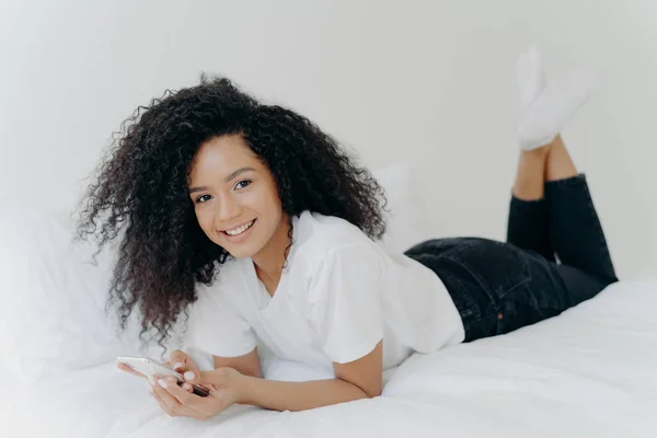 Plan intérieur d'une femme millénaire joyeuse avec coiffure afro, se couche sur le ventre dans un lit confortable, discute en ligne, vérifie la boîte e-mail, porte un t-shirt blanc, des chaussettes et un pantalon noir, a un après-midi paresseux — Photo