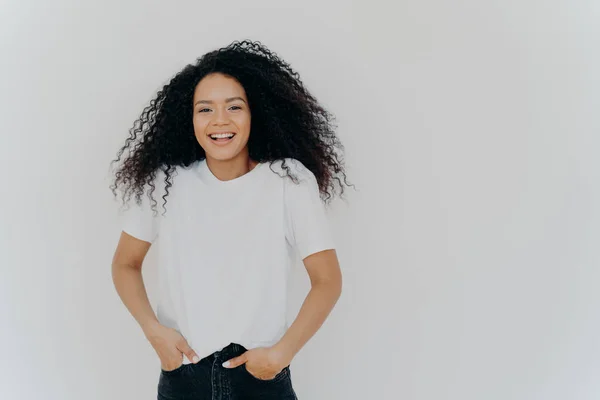 Plan à mi-longueur de belle femme souriante rit à la blague drôle, s'amuse, garde les deux mains dans des poches de jeans, porte un t-shirt blanc, a les cheveux bouclés moelleux, pose à l'intérieur, espace vide sur le côté droit — Photo