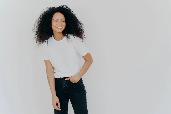 Joven mujer de pelo rizado tiene figura delgada, lleva camiseta blanca y pantalones vaqueros, mira a un lado con expresión feliz, muestra dientes sanos, nota escena divertida en el lado derecho, aislado sobre fondo blanco — Foto de Stock