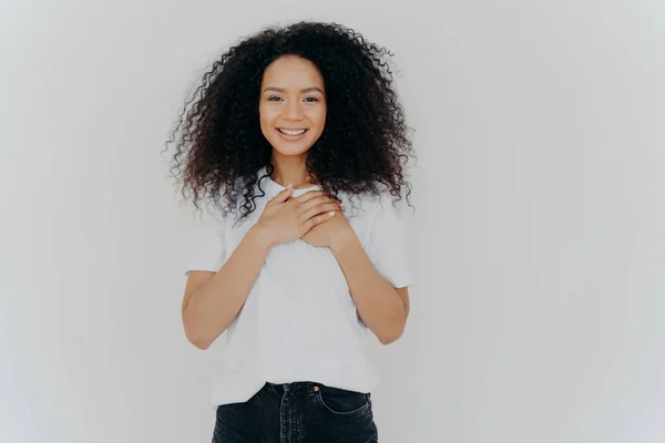 Foto de mujer alegre con pelo afro, mantiene las manos en el pecho, expresa gratitud, sonríe suavemente, usa camiseta blanca y jeans, impresionado para obtener un cumplido agradable, aislado sobre fondo blanco — Foto de Stock