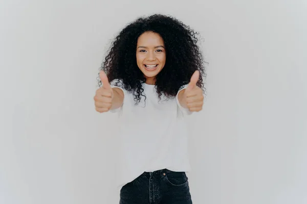 Jeune femme heureuse avec la coiffure afro, montre pouces levés, accepte ou accepte plan génial, donne son approbation, sourit largement, se tient à l'intérieur, dit excellente décision, isolé sur fond blanc — Photo