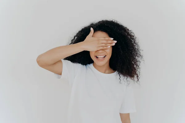 Jeune fille adolescente couvre les yeux avec la paume, se cache, sourit toothy, a Afro coiffure touffue, porte un t-shirt blanc maquillé, se tient seul en studio, attend la surprise, excité par l'anticipation — Photo