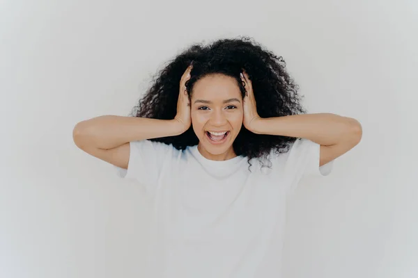 Photo de joyeuse femme à la peau foncée garde les mains sur la tête, les cheveux bouclés, rit de joie, excité par des émotions positives, vêtu d'un t-shirt blanc, entend une blague drôle, rit sincèrement. Les gens, les émotions — Photo