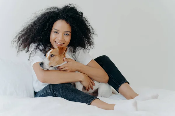 Photo de charmante jeune propriétaire d'animaux de compagnie pose dans une chambre blanche propre, étreint le chien, joue avec le meilleur ami, porte des vêtements décontractés, a une expression joyeuse. Personnes, animaux, amour, concept d'amitié — Photo