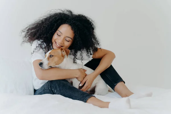 Pretty woman with curly hairstyle plays and enjoys with adorable small dog in bed, has good mood, pose together in cozy bedroom. Female wakes up with best friend. Lovely pet with owner indoor — Stockfoto