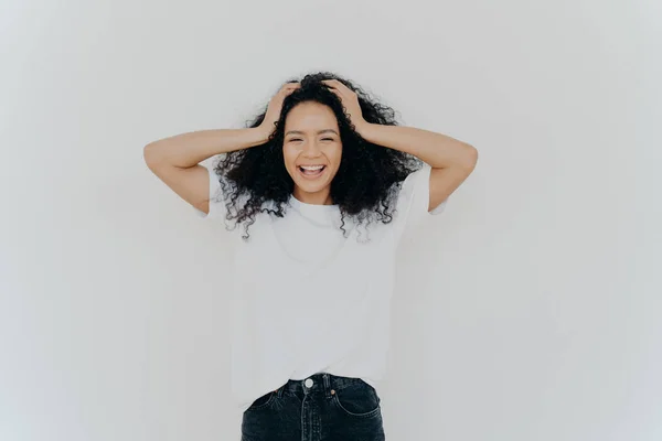 Foto av överlycklig lockigt hår kvinna skrattar glatt, har kul, klädd i vit t skjorta och jeans, ler brett, isolerad över vit bakgrund. Människor, känslor, lycka, etnicitet koncept — Stockfoto