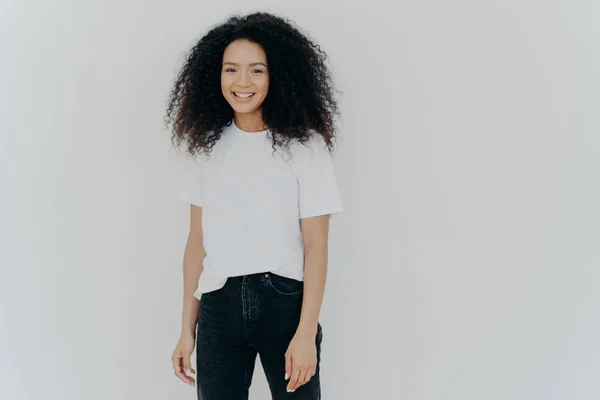 Image de belle femme avec un sourire doux, se tient satisfait, a une expression insouciante sincère, une humeur décontractée détendue, vêtu d'un t-shirt décontracté et d'un jean noir, isolé sur fond blanc — Photo