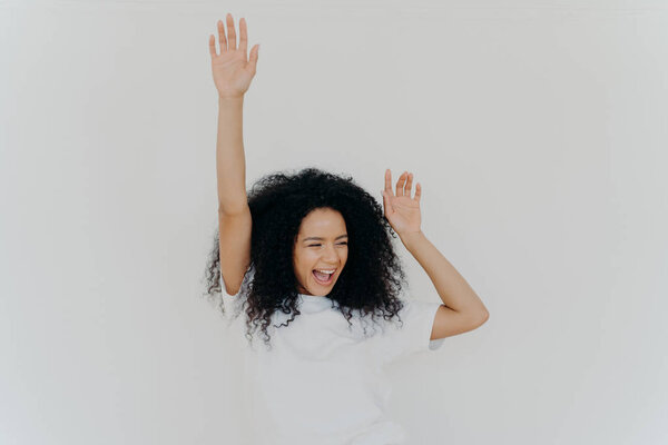 Joyful curly African American woman raises hands up, laughs from happiness, celebrates triumph, rejoices success, wears white t shirt, poses indoor, gestures actively, gets unexpected surprise