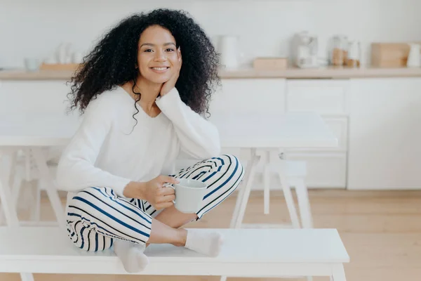 Indoor shot of pretty African American woman wears white jumper, striped pants, socks, poses on bench with cup of tea spends leisure at home in cozy kitchen feels relaxed. Female enjoys morning coffee — стокове фото