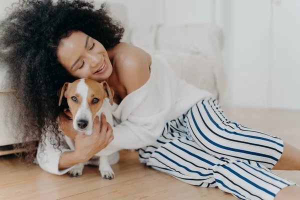 Affectionate young woman hugs dog with love and care, keeps eyes closed from pleasure, smiles gently, has healthy dark skin, poses on floor, petting animal. People, friendship and pets concept