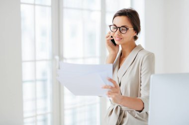 Resmi giyinmiş pozitif bir kadın, iş ortağını arıyor, finansal başlangıç projesini tartışıyor, gazetelerden bilgi okuyor, optik gözlük takıyor, ofisinde duruyor, uluslararası şirkette çalışıyor.