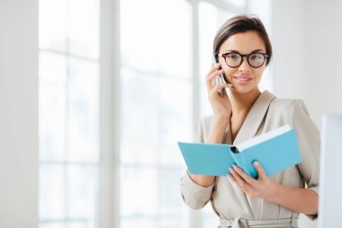 Koyu renk saçlı çekici bir kadının fotoğrafı, telefon görüşmesi, iş arkadaşıyla buluşmayı kabul etmesi, açık günlük tutması, gözlük takması ve resmi takım elbise giymesi, beyaz arka plan planları üzerine günlük poz vermesi.