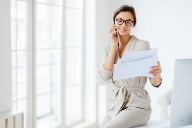 Çekici iş kadını gazete ya da iş belgeleri okur, iş ortağıyla telefonda konuşur, toplantıdan önce evrakları inceler, masaüstünde oturur, mutlu bir şekilde gülümser, belgelerle çalışır.