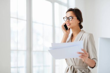Düşünceli iş kadını kameraya yan bakıyor, kağıt belgeler tutuyor, telefonla konuşuyor, mali rapor hazırlıyor, birlikte çalışma alanında poz veriyor, optik gözlük takıyor ve resmi takım elbise giyiyor. Belgeler..