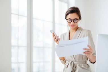 Belgelere odaklanmış, hazırlanmış raporları dikkatle okuyan, modern akıllı telefonları olan, şeffaf gözlükler takan ve ofis içi çeklerin üzerine poz veren kadın çalışanlar.