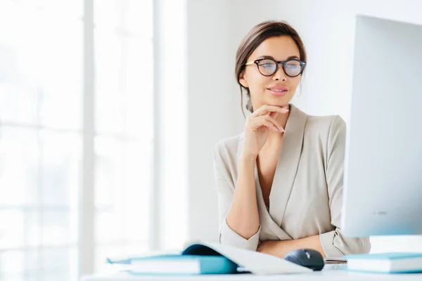 Charming female office worker keeps hand under chin, looks in screen of computer, wears spectacles for vision correction, poses in own cabinet, prepares information for future business meeting