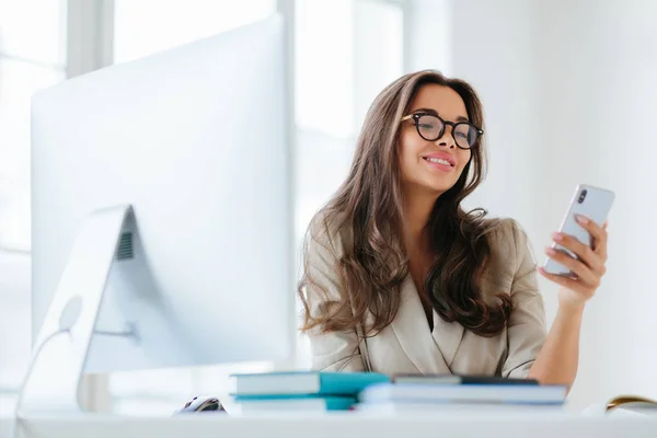 Smiling female in corporate clothes watches funny video via smartphone, sits at desktop in office, uses computer for work, enjoys working process, browses information for project, wears eyeglasses