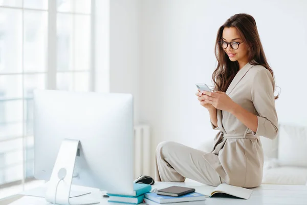ベージュの公式スーツのエレガントな女性は、ウェブサイト上のニュースを注意深く読み取り、キャビネット、コンピュータモニタとノートパッドだけでデスクトップに座って周り。女性社員はオフィスで現代的なスマートフォンを使う — ストック写真