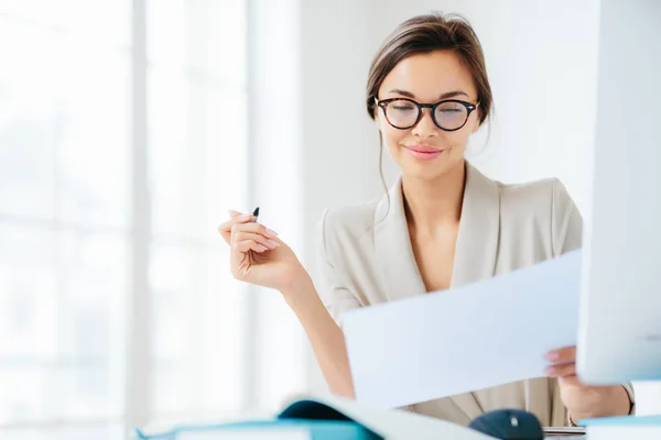 La donna d'affari di successo concentrata guarda attentamente in carta, studia termini di contratto, tiene la penna, scrive in documentazioni, vestito formalmente, posa a desktop contro interno spazioso bianco — Foto Stock