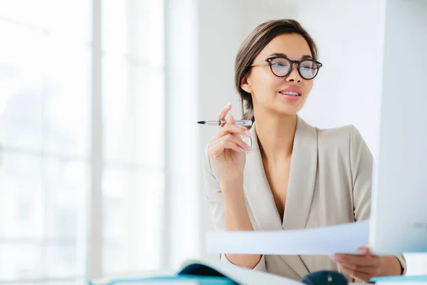 Framgångsrik affärskvinna inriktad på bildskärm av datorn, studier ekonomiska frågor, innehar dokument och penna, arbetar på kontoret, bär formella kläder och glasögon, poser över vit bakgrund. — Stockfoto