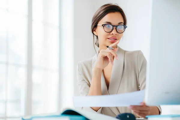 Economista femminile posa nello spazio di coworking, focalizzata nel monitorare, verifica o controlla le cifre dal rapporto finanziario, detiene documento cartaceo e penna, ha i capelli pettinati scuri, indossa abito da lavoro formale — Foto Stock