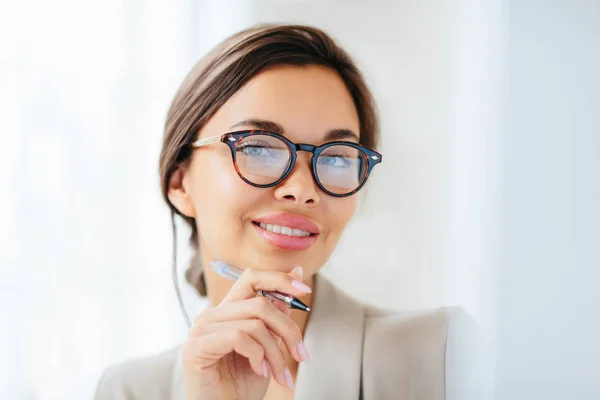 Agradável aparência jovem modelo feminino com maquiagem e manicure segura caneta, toca o queixo suavemente, olha diretamente para a câmera, vestida com roupas formais, trabalha no escritório da empresa, usa óculos ópticos — Fotografia de Stock