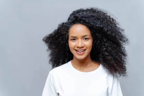 Positieve krullende vrouw met natuurlijke schoonheid, gekleed in wit casual t-shirt, heeft een vrolijke uitdrukking, kijkt direct naar de camera, poseert tegen een grijze achtergrond. Vrolijk tienermeisje drukt goede emoties uit — Stockfoto