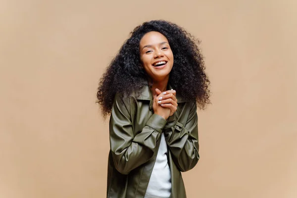 Positif jolie femme afro-américaine a les cheveux bouclés, garde les mains ensemble, heureux de rencontrer un ami, habillé en vêtements à la mode, sourit largement, modèles sur fond brun. Wow, c'est sympa. — Photo