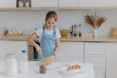 Şirin küçük kız çizgili önlük giyiyor, malzemeleri kaseye çırpıyor, hamur hazırlıyor, yemek yapmayı öğretiyor, yumurta, süt ve un ile mutfak arka planında beyaz masada duruyor. Childern ve aşçılık konsepti