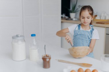 Çocuklar, yemek pişirme ve ev konsepti. Güzel, siyah saçlı bir kız kasede malzeme çırpıyor, pastaya hamur hazırlamakla meşgul, geleceğin aşçısı oluyor, beyaz tişört ve çizgili önlük giyiyor,