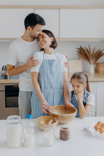 Felice bella famiglia in cucina di casa, padre abbraccia la madre con amore, bambina guarda in ciotola, osserva come la mamma cucina e frusta gli ingredienti, utilizzare le uova per fare la pasta. Atmosfera domestica — Foto Stock