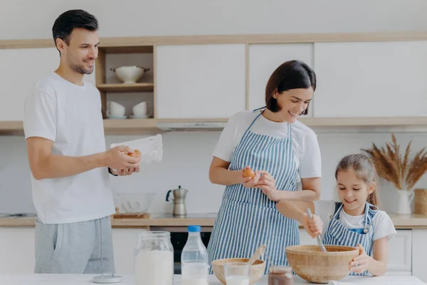 Happy parents cook together with daughter, pose at modern home kitchen give eggs to add to dough, small kid whisks ingredients in bowl have glad expressions. Food and family concept. Cooking breakfast