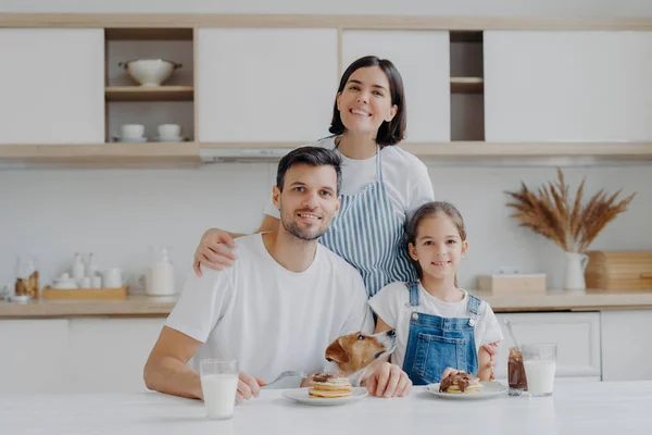 Ritratto di famiglia di madre felice, figlia e padre posano in cucina durante l'ora di colazione, mangiano deliziose frittelle fatte in casa, il loro cane posa vicino, hanno buone relazioni amichevoli, si amano — Foto Stock