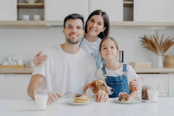 Famiglia, tempo libero, passatempo, concetto di mangiare. Padre, madre e figlia, Jack Russell terrier cane posare tutti insieme alla fotocamera contro l'interno della cucina, ottenere il piacere di mangiare frittelle bere latte fresco — Foto Stock