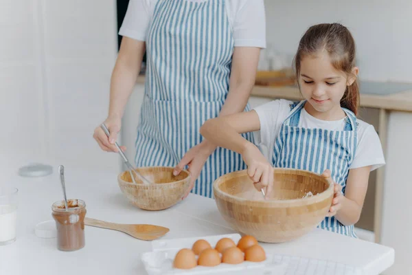 Mother gives culinary lesson to little child, stand next to each other, mix ingredient in big wooden bowls, make dough together, use eggs, flour and other products. Family, cooking, motherhood