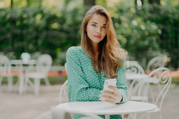 Tiro ao ar livre da mulher na moda com cabelo longo, vestido com camisa polkadot verde, usa celular moderno para a comunicação, conectado à internet, senta-se à mesa branca no café exterior, espera por ordem — Fotografia de Stock