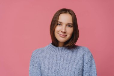 Hoş görünümlü, koyu renk saçlı, örgü kazaklı bir kadının yatay görüntüsü kameraya şefkatle bakıyor, kış günlerinden ve sıcacık havadan hoşlanıyor, pembe arka planda poz veriyor. Kadınlık ve duygular kavramı