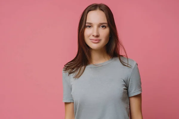 stock image Adorable millennial girl with straight dark hair, wears casual t shirt, looks seriously at camera, has healthy skin, poses over pink background. Pleasant looking woman has talk with someone.